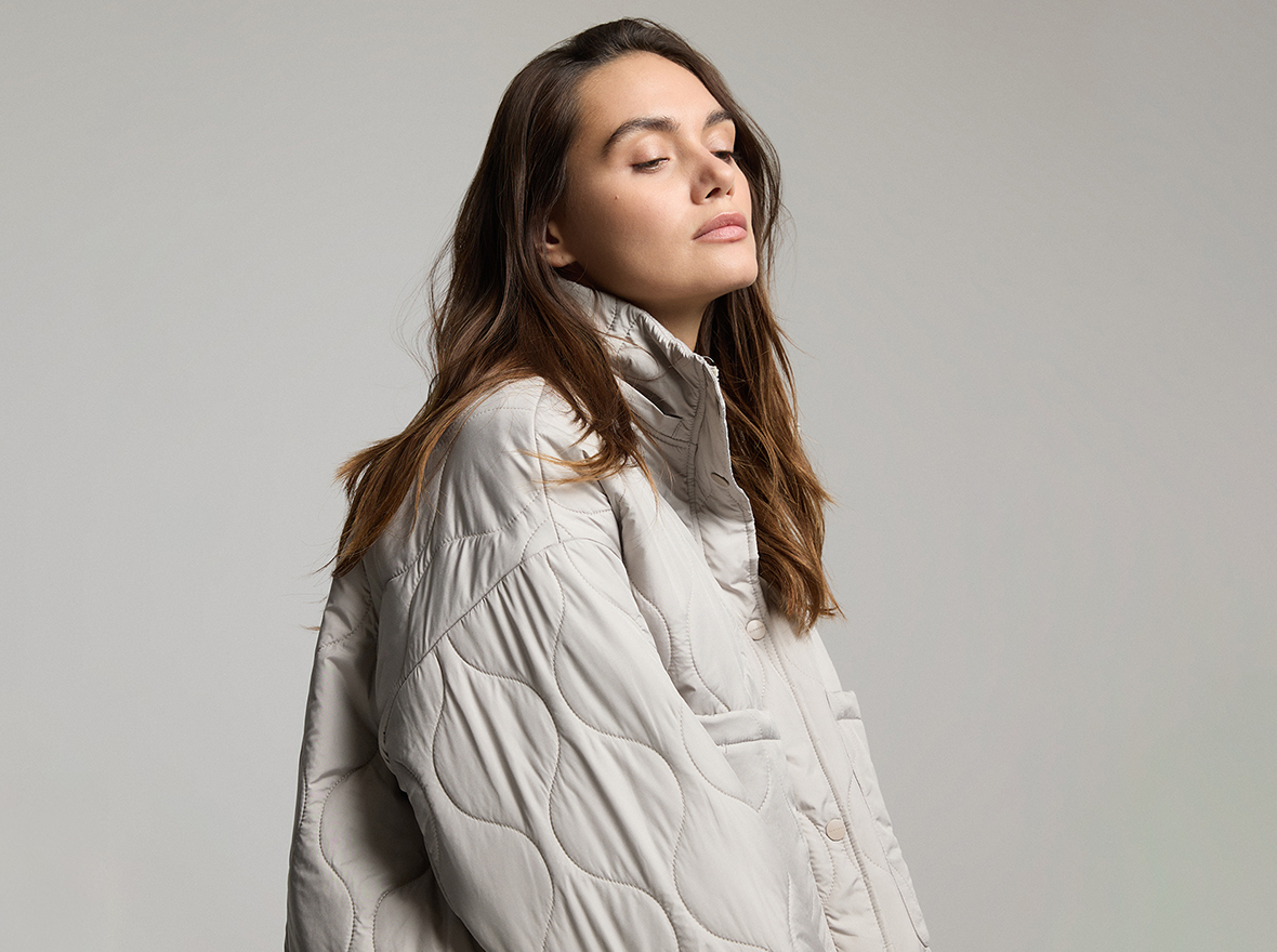  Młoda modelka siedząca w studio, ubrana w jasną kurtkę. Ujęcie wykonane z lekkiego profilu, tworzące delikatną i elegancką atmosferę.